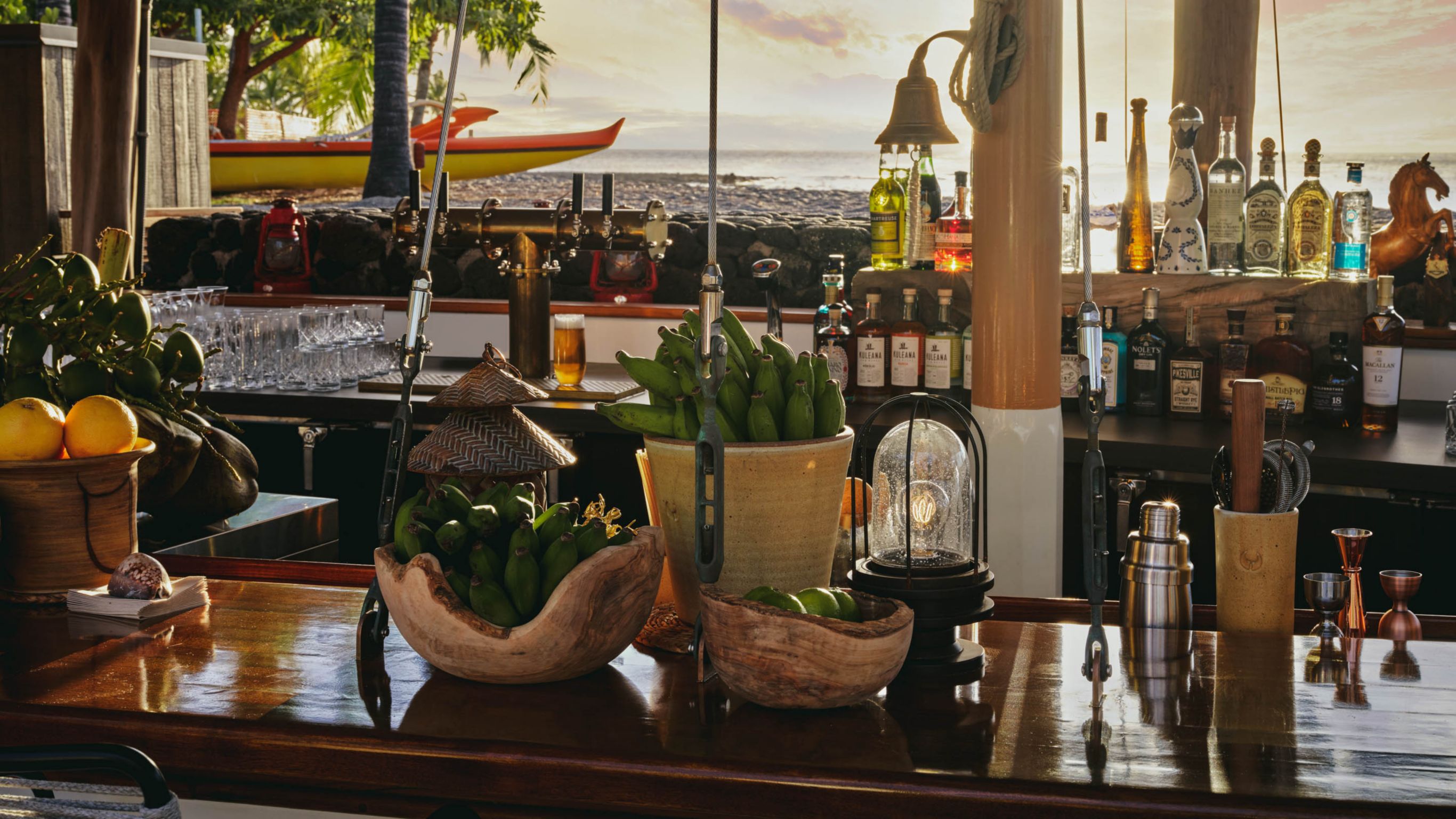 A beachside bar with a wooden counter, featuring bowls of fruit, bottles, and a view of the ocean at sunset.A beachside bar with a wooden counter, featuring bowls of fruit, bottles, and a view of the ocean at sunset.